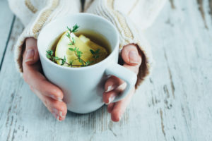 Two hands hold tea on wooden table. Tea has thyme leaves and lemon slices in it