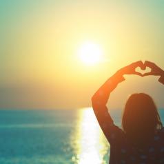 Rear view of person standing by beach looking out to sea with hands forming heart shape